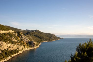 Güneşin doğuşundaki yeşil kıyı manzarasının büyüleyici bir görüntüsü, dalgalı tepeler ve sakin sular tarafından çerçevelenmiş, sükuneti ve doğanın sabahki güzelliğini somutlaştıran.