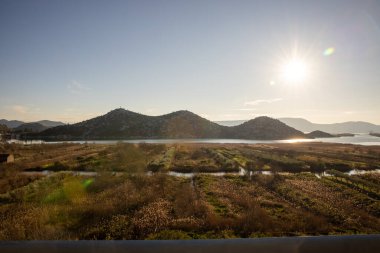 Güneş ışınları, sakin sulara yansıyan yuvarlanan tepeleri aydınlatır. Bu resim gibi manzarada ışık ve doğa arasında büyüleyici bir uyum yaratır..