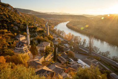 A breathtaking view of a picturesque village alongside a winding river, framed by lush hills, illuminated by the warm light of the setting sun at golden hour. clipart