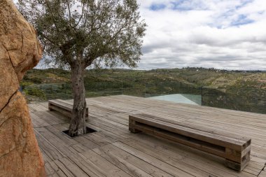 Ahşap bir güverteyi doğal bir ortamda süsleyen yalnız bir ağaç, geniş bir tepe manzarası sunuyor ve bulutlu bir gökyüzü, doğayla birleşen bir tasarım..