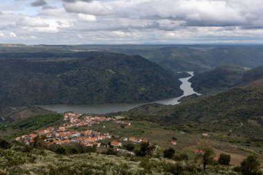 Çarpıcı bulutlu bir gökyüzünün altında kıvrımlı bir nehirle çevrili kırmızı çatılı evleri olan büyüleyici bir köy..