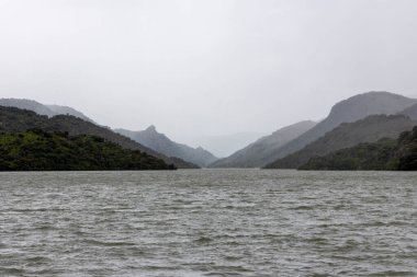 Bulutlu bir günde, yemyeşil tepelerle çevrili bir vadiden akan sakin bir nehir huzur ve doğanın güzelliğini çağrıştırıyor..