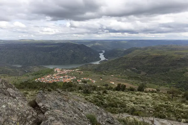 Resimli bir köy, yemyeşil tepelerin arasında kıvrılan bir nehrin kenarında, dramatik bulutlu bir gökyüzünün altında, ahenkli ve sakin bir manzara yaratıyor..
