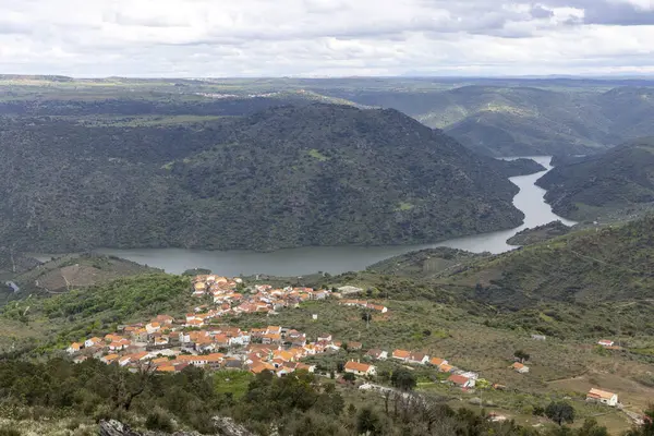 Bulutlu bir gökyüzünün altında yemyeşil bir arazide, dingin bir nehrin kenarına kurulmuş kırmızı çatıları olan pitoresk bir köy, nefes kesici manzaralar sunuyor..