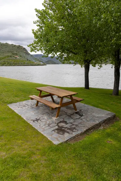 Sakin bir gölün yanındaki taştan bir platformda duran ahşap piknik masası yemyeşil çimenler ve bulutlu bir gökyüzünün altındaki uzak tepelerle çevrilidir..