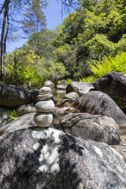 Çerçevenin merkezinde dikkatlice dengelenmiş gri ve beyaz taş yığınını gösteren yakın plan bir çekim. Taşlar daha büyük, daha koyu gri nehir kayalarının arasına yerleşmiş.. 