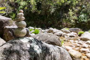 Sığ bir alan derinliği, dikkatle dengelenmiş nehir kayaları yığınını tasvir eder. Cairn olarak da bilinir. Daha büyük, liken kaplı bir kayanın üzerine yerleştirilmiştir..