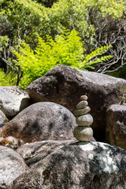 Görüntü, çeşitli boyutlardaki birkaç kayanın yakın çekimini gösteriyor. Ön planda, daha büyük ve düzensiz şekilli gri kaya parçalarına karşı belirgin bir şekilde dengelenmiş küçük bir çakıl taşı cairn 'i bulunur..