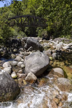 Nehrin sığ bir bölgesini gösteren yüksek açılı, yakın çekim. Ön planda hem pürüzsüz hem de pürüzsüz dokulara sahip çeşitli gri ve açık gri nehir kayaları bulunmaktadır..