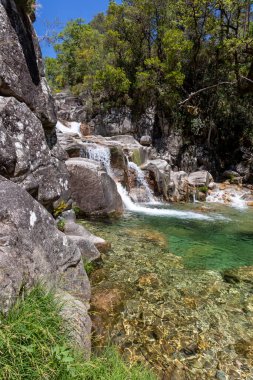 Görüntü, küçük bir şelalenin, gri ve açık gri kayalardan oluşan bir kümenin üzerine dökülen temiz ve sığ bir su havuzunun yakın görüntüsünü sergiliyor. Su pürüzsüz, akıcı bir doku sergiler..