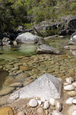 Yüksek açılı, yakın plan, sığ bir nehir yatağını tasvir ediyor. Görüntü, ağırlıklı olarak açık kahverengi ve bej renklerinde kristal berraklığında, sığ sulara batmış pürüzsüz, değişken taşlardan oluşan bir koleksiyon sergiliyor..