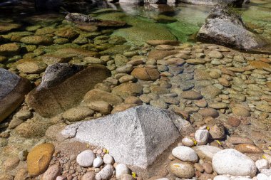 Yüksek açı, yakın çekim, sığ bir su kütlesi ve çeşitli boyutlarda pürüzsüz yuvarlak taşlardan oluşan bir nehir yatağı tasvir ediyor. Su fevkalade berrak, su altındaki kayaların ve dokularının detaylı bir görüntüsünü sağlıyor..