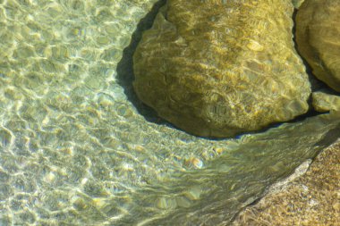 Yüksek açılı, yakın plan, yüzeydeki su yüzeyini su altında kalan kayalarla tasvir ediyor. Su ince dalgalanmalar gösterir, ışık kırılmasına neden olur ve suyun yüzeyinde dokulu bir desen oluşturur..