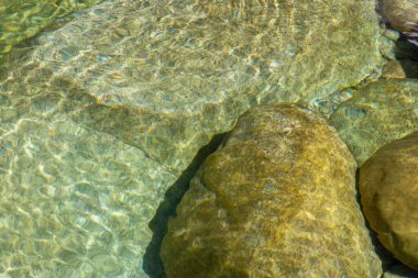 Birkaç kayanın yakın çekim görüntüsü kısmen temiz suya batmış. Görüntü, ışık ve suyun etkileşimini gösteriyor, bu da kayaların yüzeyinde görünür ışık kırılması ve dalgalanma desenlerine neden oluyor..