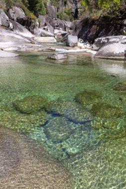 Yüksek açılı, yakın çekim. Sığ bir su kütlesi ve görülebilir kayalık bir tabanı gösteriyor. Su son derece berrak, yeşilimsi bir renk sergiliyor. Değişen büyüklükteki su altı kayaları dibe saçılmış..