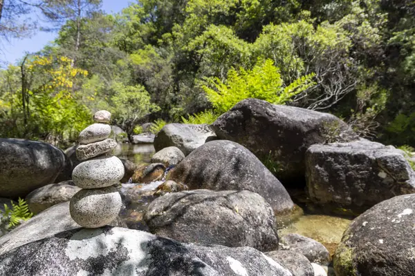 Görüntü titizlikle dengelenmiş gri ve açık kahverengi taş yığınını küçük bir cairn oluşturarak sergiliyor. Bu cairn daha büyük, daha koyu gri bir kayanın üzerinde sığ bir dere ya da nehir kenarında yer alır..