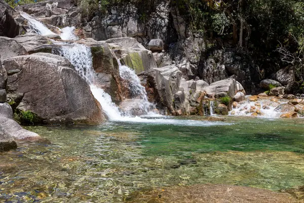 Görüntü, kayaların üzerinden akan çağlayan bir şelalenin sığ bir su havuzuna aktığını gösteriyor. Su, değişen derecede opaklığa sahip pürüzsüz, akıcı bir doku sergiler. Kayalar ağırlıklı olarak gri ve kahverengi..