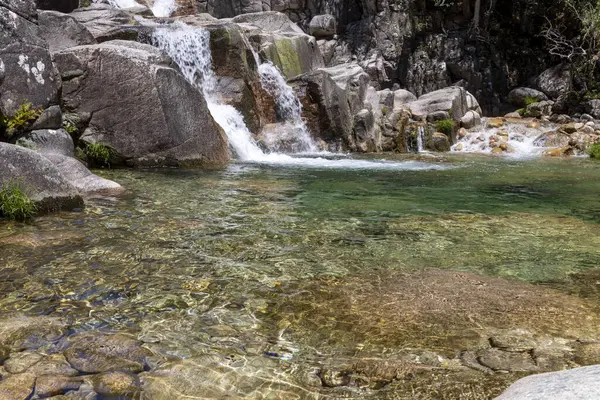 Görüntü, küçük bir şelalenin sığ ve kayalık bir havuza dökülüşünün yüksek açılı ve yakın görüntüsünü sergiliyor. Su son derece berraktır, yüzeyin altındaki kayaların dokusunu ve su bitkilerini ortaya çıkarır..
