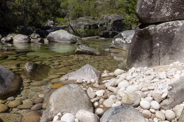 Görüntü yüksek açılı, sığ bir yatağın yakın çekim görüntüsünü sergiliyor. Su berrak, akarsu yatağındaki taşları ve çakıl taşlarını ortaya çıkarıyor..