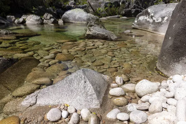 Görüntü yüksek açılı, sığ bir dere yatağının yakın görüntüsünü veriyor. Dere yatağı ağırlıklı olarak çeşitli kaya ve çakıl taşlarından oluşur, küçük, pürüzsüz taşlardan büyük, açısal kayalara kadar uzanır..