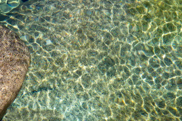 Yüksek açılı, yakın plan, yüzeydeki pürüzsüz, yuvarlak çakıl taşlarından oluşan yatağın üzerindeki berrak suyu tasvir ediyor. Su, ince dalgalanmalar ve ışık kırıntıları sergiliyor. Çerçevede dokulu bir desen oluşturuyor.. 