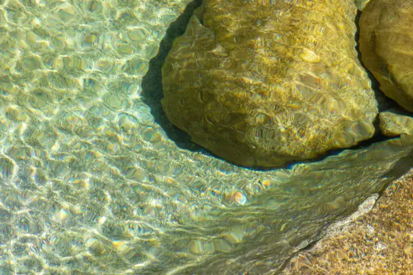 Yüksek açılı, yakın plan, yüzeydeki su yüzeyini su altında kalan kayalarla tasvir ediyor. Su ince dalgalanmalar gösterir, ışık kırılmasına neden olur ve suyun yüzeyinde dokulu bir desen oluşturur..