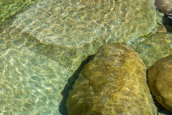 Birkaç kayanın yakın çekim görüntüsü kısmen temiz suya batmış. Görüntü, ışık ve suyun etkileşimini gösteriyor, bu da kayaların yüzeyinde görünür ışık kırılması ve dalgalanma desenlerine neden oluyor..