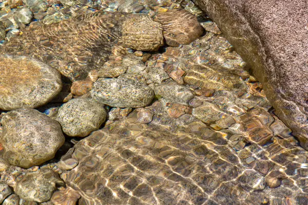 Yüksek açılı, yakın çekim, birkaç pürüzsüz, yuvarlak kayanın yüzeysel ve berrak suya batışını gösteriyor. Suyun yüzeyi, kayaların ve nehir yatağının altında ışık kırılması ve bozulmasına yol açan görünür bir dalga etkisi gösteriyor.. 