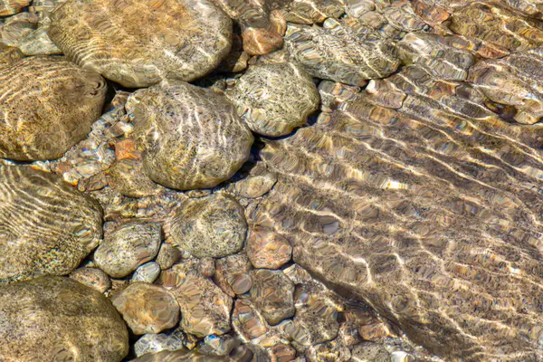Nehir yatağının dokusunu gösteren yüksek çözünürlüklü yakın plan görüntü. Yüzeysel ve berrak suyun altında çeşitli boyutlarda sayısız pürüzsüz ve yuvarlak taşlar görülebilir..