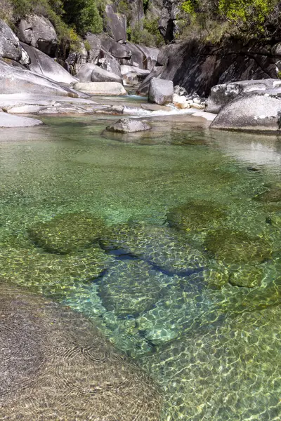 Yüksek açılı, yakın çekim. Sığ bir su kütlesi ve görülebilir kayalık bir tabanı gösteriyor. Su son derece berrak, yeşilimsi bir renk sergiliyor. Değişen büyüklükteki su altı kayaları dibe saçılmış..