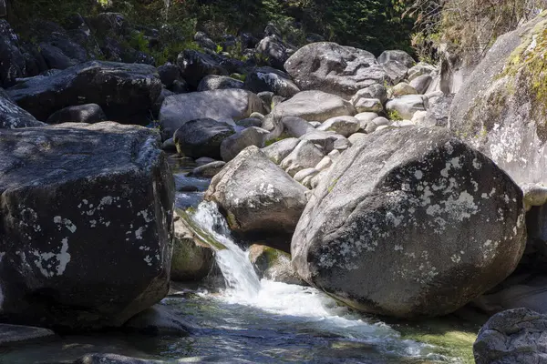 Yüksek açılı, geniş, koyu gri ve desenli kayalar arasında akan küçük bir akarsuyun yakın görüntüsü. Su nispeten berrak görünüyor ve hareket bulanıklığı gösteriyor..  