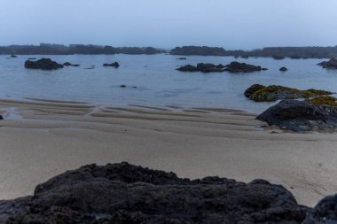 Şafak veya gün doğumunda sakin bir sahneyi resmeden panoramik bir manzara. Görüntüde ön planda kumlu bir sahil var. Kumdan koyu kayalar fışkırıyor..  
