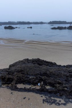 Yüksek açılı, bulutlu bir gökyüzünün altında ufka uzanan kumlu bir plaj görüntüsü. Kum açık renklidir ve ince dokusal varyasyonlar gösterir. Koyu renkli kayalarda belirgin bir özellik vardır..