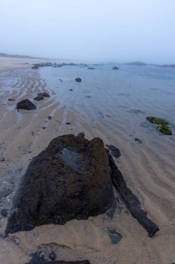 Yüksek açı, alçak gelgitte kumlu bir sahilin yakın görüntüsü. Görüntü koyu renkli kayaların sığ ve berrak sulara kısmen battığını gösteriyor. Kum ince dalgalanma desenleri sergiliyor..