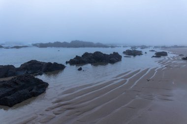 Görüntü alçak açılı, kıyı sahnesinin uzak bir görüntüsünü gösteriyor. Karanlık, ıslak kayalar kıyıya yakın sığ ve sakin sularda kısmen su altında kalır. Ön plandaki kum farklı gelgit izleri gösteriyor.. 