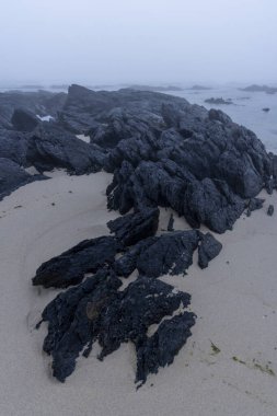 Yüksek açılı, koyu renkli volkanik kayaların yakın görüntüsü açık bej renkli kumlu bir yüzeye saçılmış. Kayalar değişik boyutlarda ve şekillerde pürüzlü ve düzensiz bir doku sergilerler..