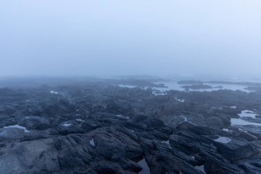 Yüksek açılı, yoğun bir sis veya siste gizlenmiş kayalık bir sahil şeridi görüntüsü. Renk paleti ağırlıklı olarak gri ve mavilerden oluşur. Kayalar koyu ve pürüzlü görünüyor..