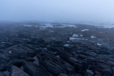 Görüntü, sis veya sisle kaplanmış kayalık bir arazinin geniş bir görüntüsünü gösteriyor. Genel renk paleti sessiz maviler ve gri renktedir. Ön planda koyu, pürüzlü kayalar var..