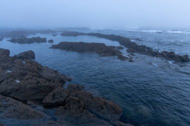 Görüntü, sakin, koyu mavi bir okyanusa kısmen batmış karanlık ve düzensiz kayaların hakim olduğu bir kıyı sahnesini tasvir ediyor. Kalın bir sis tabakası arkaplanı gizler ve görünmezliği azaltır..