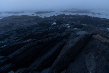 Yüksek açılı, şafakta ya da gün doğumunda kıyı kayalıkları görüntüsü. Sahne sisli ve sisli bir ortamda karamsar bir ortam yaratıyor. Kayalar koyu gri ve siyah..