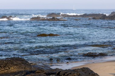 Görüntü, kayalık bir kıyı şeridine çarpan okyanus dalgalarına odaklanmış bir deniz manzarasını gösteriyor. Renk paleti ağırlıklı olarak mavi ve turkuazdır, gölge çeşitleri derinliği temsil eder.