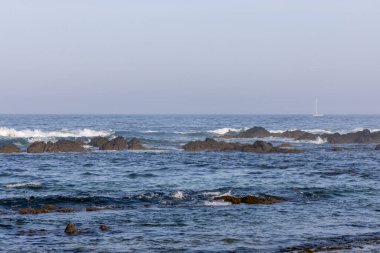 Görüntü kıyı sahnesinin uzak bir görüntüsünü gösteriyor. Ön planda kısmen mavi-gri sulara gömülü koyu renkli kayalar ve onlara doğru gelen küçük dalgalar var.. 