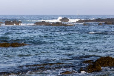 Görüntü, kıyı boyunca uzanan karanlık kayalara çarpan dalgalı bir su kütlesinin geniş bir görüntüsünü gösteriyor. Su ağırlıklı olarak mavi ve turkuazdır. Dalgaların olduğu yerde beyazlar görülebilir.