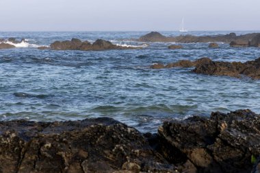 Görüntü, okyanus dalgalarıyla dolu kayalık bir kıyı şeridini betimliyor. Ön planda koyu renkli kayaların kısmen suya battığını gösteriyor. Orta yerde su yüzeyinde hareket eden küçük dalgalar var.