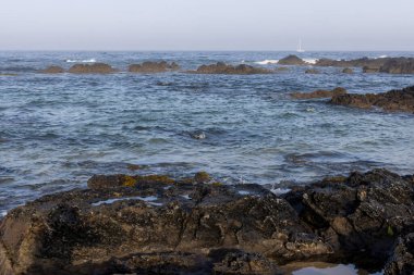 Görüntü, ön planda koyu renkli kayaların olduğu bir sahneyi gösteriyor. Orta zemin ve arka plan görünür dalgaları ve biraz pürüzlü dokusu olan bir su kütlesine sahiptir..