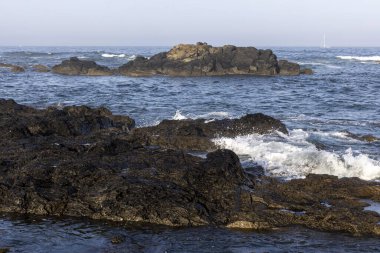 Görüntü, sudan çıkan sert kayaların olduğu bir kıyı sahnesini gösteriyor. Okyanus dalgaları görülebilir, kayalara çarptıkları yerde beyaz köpük görülebilir. Su koyu mavi..
