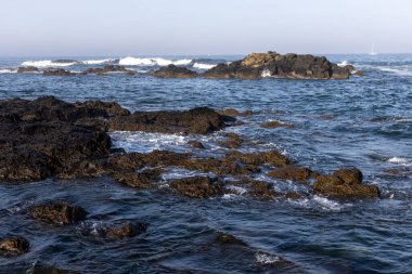 Görüntü, okyanusa kısmen batmış koyu dokulu kayaların hakim olduğu bir kıyı sahnesini gösteriyor. Küçük dalgalar görülebilir, suyun üzerinde dokulu bir yüzey oluşturur. Su koyu mavi görünür.