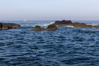 Görüntü, okyanus dalgalarıyla kayalık bir sahil şeridini gösteriyor. Su koyu mavi renktedir, dalgalarda beyaz noktalar görülebilir. Koyu renkli kayalar kısmen suya gömülüdür..