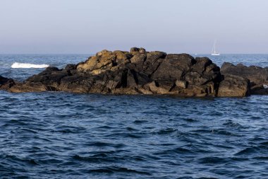 Görüntü, bir su kütlesinin ortasında koyu renkli kayalık bir çıkıntı gösteriyor. Kayalar düzensiz ve koyu gri ya da siyah gibi görünüyor..