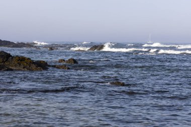 Görüntü dalgaların kayalık bir kıyı şeridine çarptığı engebeli bir denizi gösteriyor. Su ağırlıklı olarak koyu mavi ve beyaz noktalar aktif dalgaları gösteriyor.. 